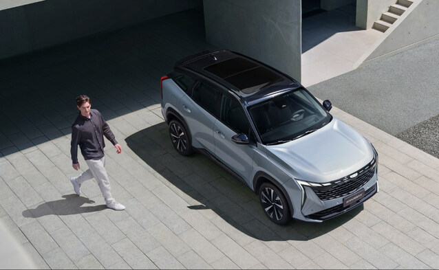 Srebrny SUV Geely z czarnym dachem zaparkowany przed nowoczesnym budynkiem, obok przechodzi mężczyzna