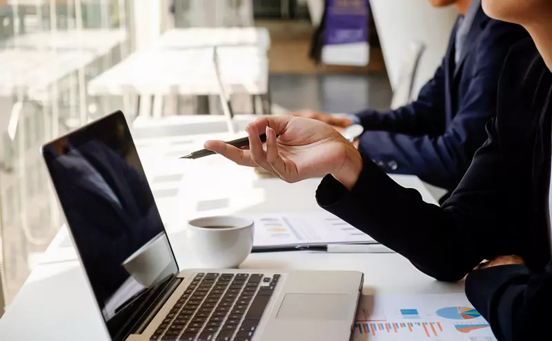 Laptop na stole w sali konferencyjnej i po prawej stronie widoczne tylko ręce 