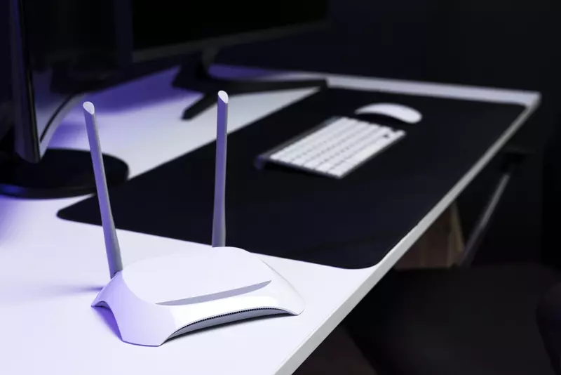 Biały router Wi-Fi z dwiema antenami na biurku, w tle monitor, klawiatura i mysz