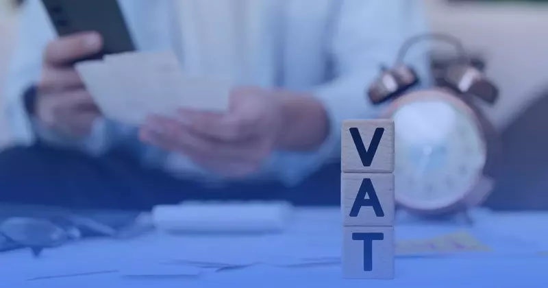 Drewniane klocki z napisem VAT na biurku, w tle mężczyzna z dokumentami i telefonem oraz budzik