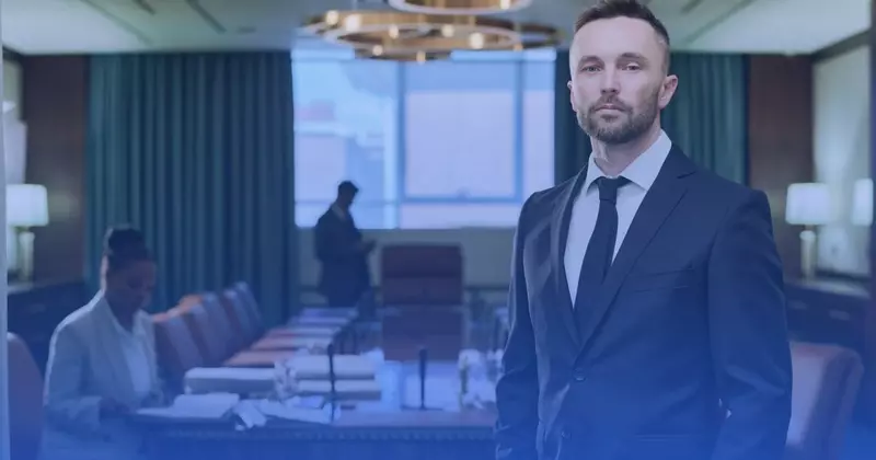Mężczyzna z brodą w granatowym garniturze i krawacie stojący w sali konferencyjnej, w tle dwie osoby przy stole
