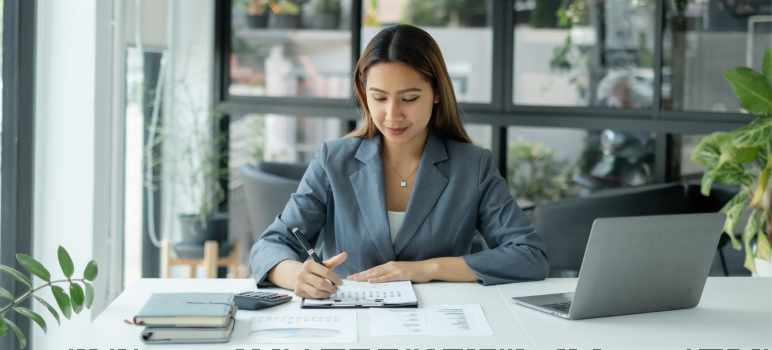 Uśmiechnięta kobieta ubrana biznesowo siedząca przy biurku z laptopem i pisząca długopisem