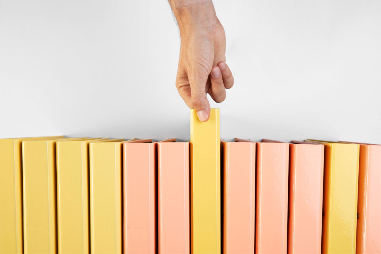 Kilka segregatorów żółtych i różowych ustawionych pionowo jedne obok drugiego i widoczna dłoń która wyciąga jeden z segregatorów